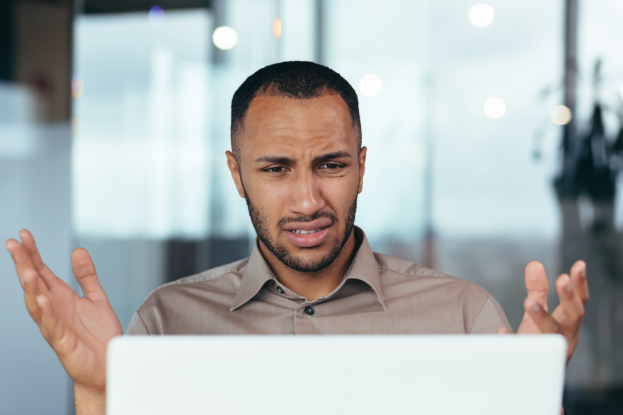 A businessman looks at his computer screen, with his hands up, confused and frustrated by dark patterns on the website he is viewing.